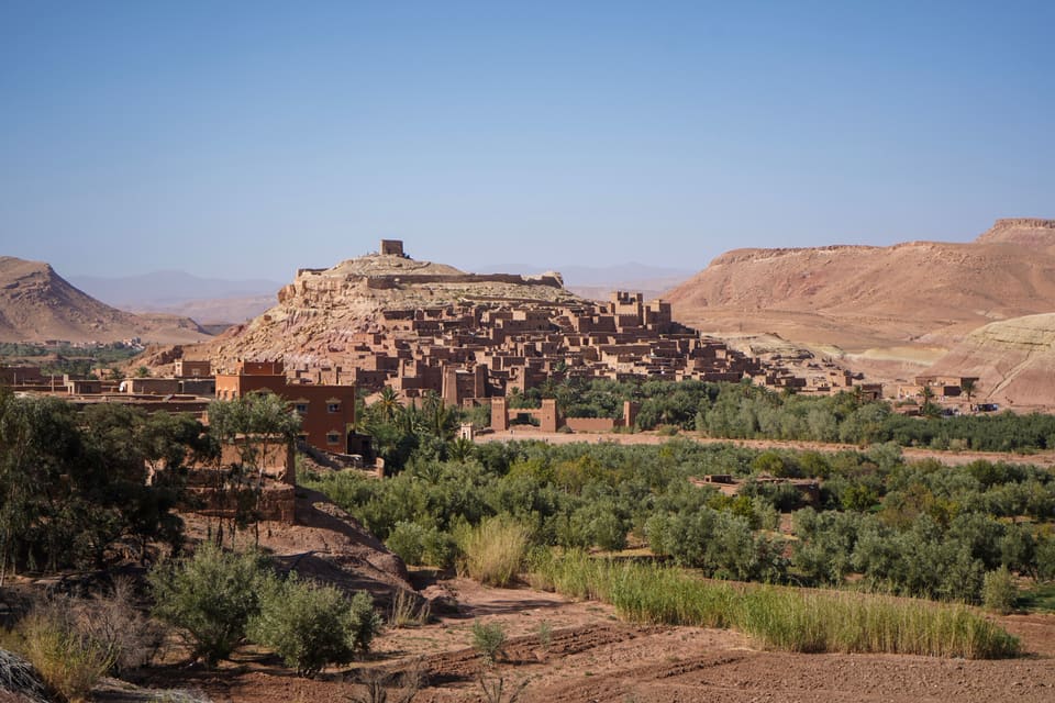 A fortified village of red earth climbing a hill to a ruined tower, above a green strip of palms and olive trees.