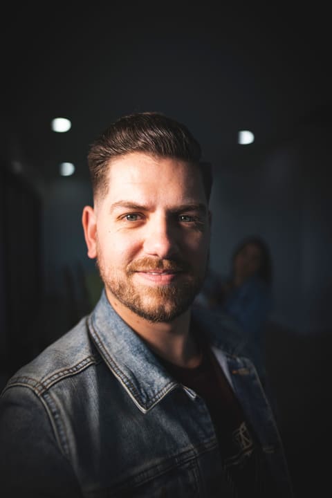 Alex Voigt: short dark hair, a trimmed beard and a half smile, in a denim jacket, lit from one side against a dark room.