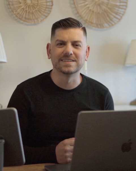 Alex Voigt at a wooden desk, looking at the camera: short dark hair, a trimmed beard, a black sweater. A laptop and an iPad on a stand are in front of him, and two woven plates hang on the white wall behind.