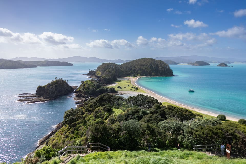 From a grassy lookout: a wooded island joined to the next by a curve of sand, with turquoise water on one side of it, deep blue on the other, and a small boat at anchor.