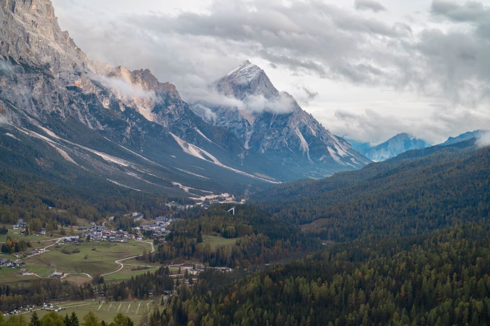 A village on a valley floor of autumn larch, under a grey rock peak with cloud caught on its shoulder.