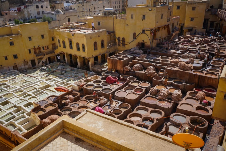 A tannery from above: rows of round stone vats of dye with hides piled on their rims, a few workers standing among them, and yellow buildings all around.