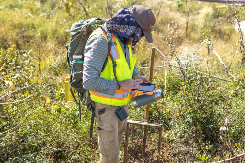A field archaeologist in a hi-vis vest and sun hat enters data on an iPad in a rugged case, standing next to a shovel and a soil screen.