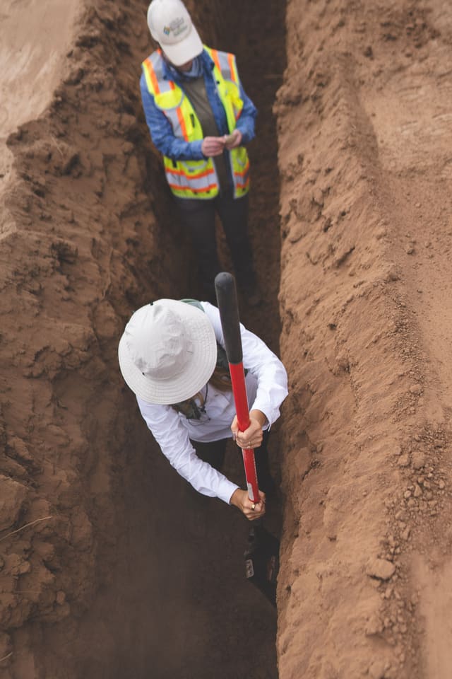 Two people in hard hats and hi-vis vests in a deep, narrow trench, seen from above. One is scraping the trench wall with a long-handled tool.