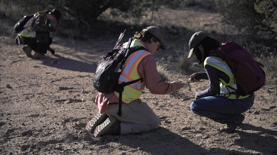 Still from the film: Three surveyors in hi-vis vests kneel on open ground, looking closely at something in the soil.