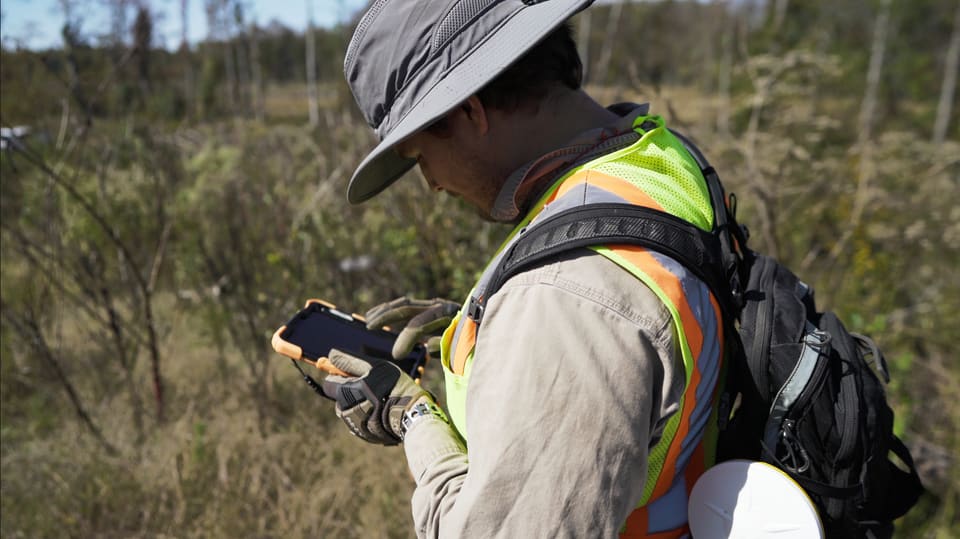 Still from the film: A surveyor in a sun hat and hi-vis vest looks down at a phone in a rugged case, in green scrub.