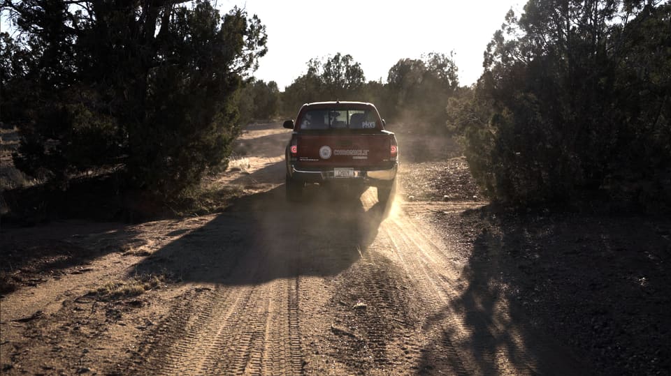 Still from the film We’re on a Mission: a pickup truck drives toward the camera down a dirt road, its dust lit from behind by a low sun.