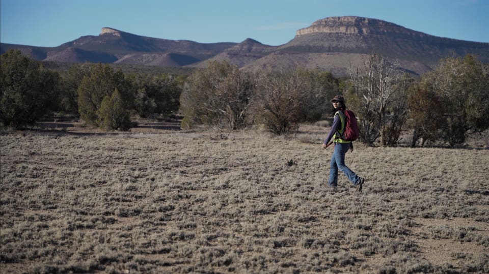 Still from the film: a surveyor in a hi-vis vest walks across open desert scrub, with a flat-topped mesa behind.