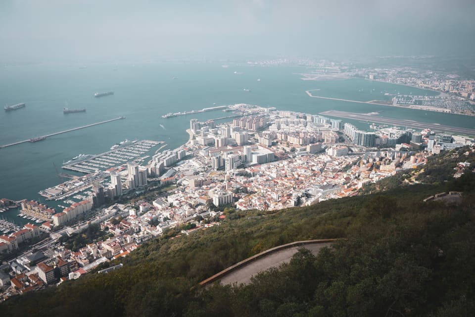 Looking down from the Rock on the town, its marinas and the airport runway, with ships at anchor in a hazy bay.