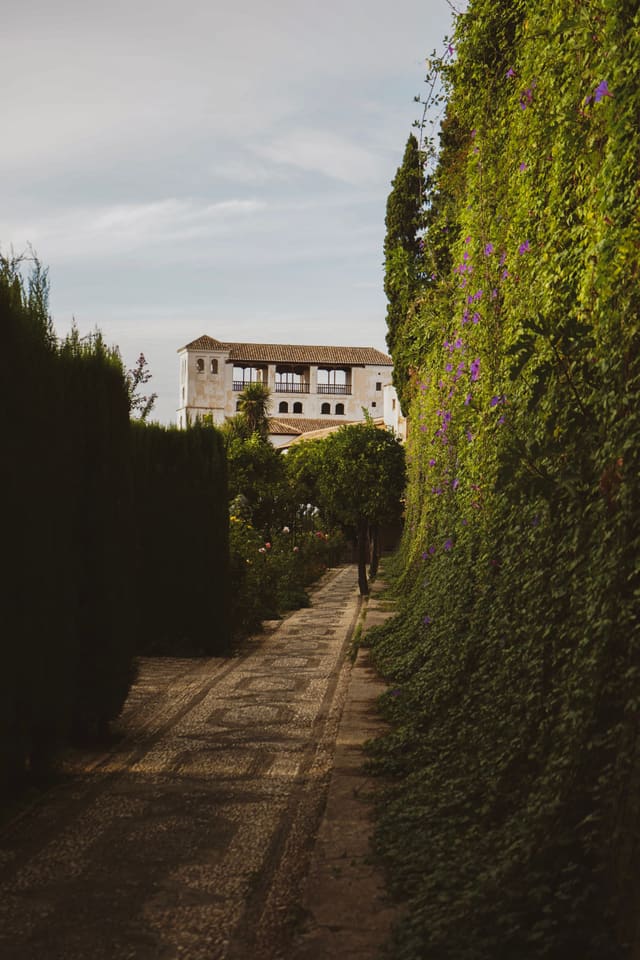 A pebble-mosaic path between a tall clipped hedge and a wall of ivy with purple flowers, leading to a white pavilion with an arcaded gallery.