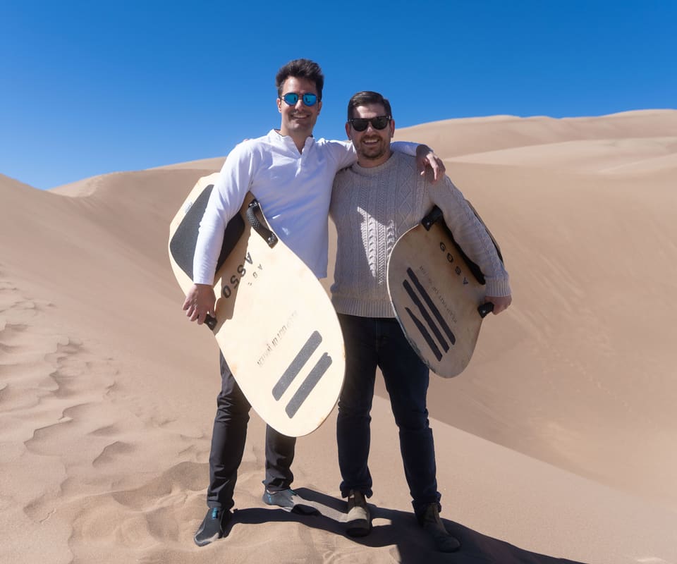 Alex and Hugo stand on a sand dune with an arm round each other’s shoulders, each holding a wooden sandboard, under a clear blue sky.