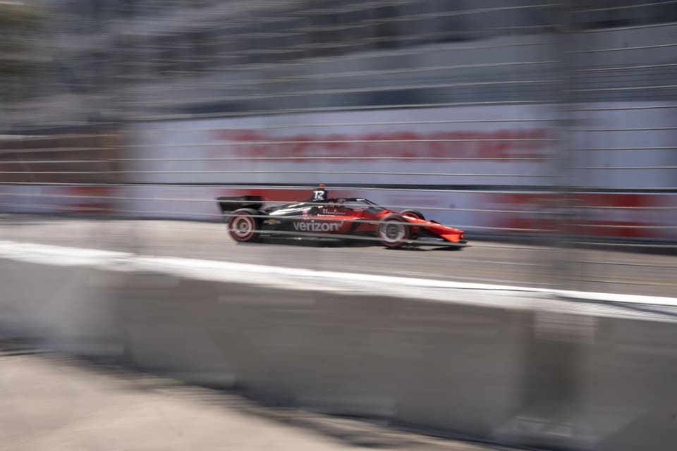 A black and red IndyCar at speed, sharp against a streaked blur of fence and wall.