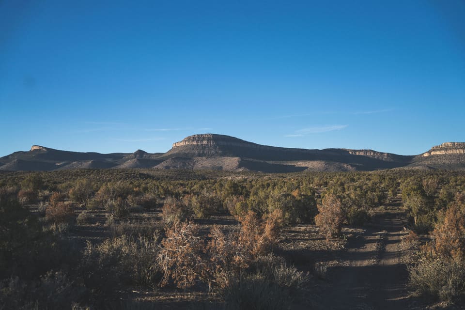 A flat-topped mesa on the horizon above dark desert scrub, under a deep blue evening sky.