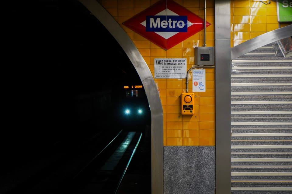 A train’s headlights coming out of a dark tunnel, beside a wall of yellow tile with the red and blue Metro sign on it.