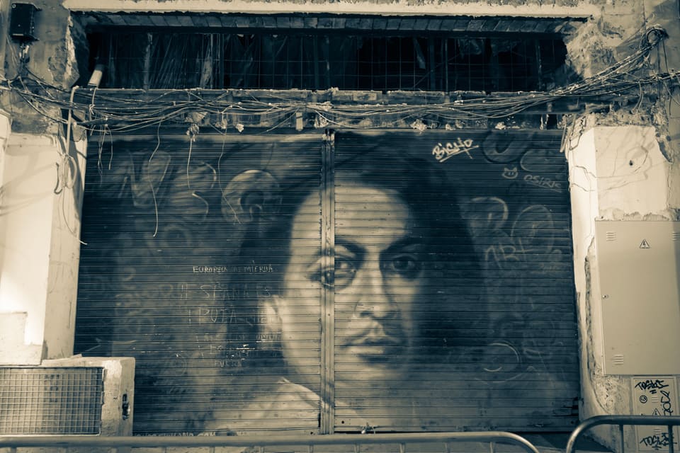 A face painted across a shop’s rolled-down shutter, in the manner of an old portrait, with graffiti written over it and loose wiring strung above.
