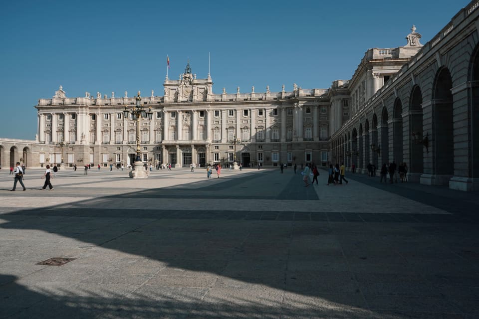 The palace’s long stone front across a wide paved courtyard, half of it in the shadow of an arcade, with a few people walking through.