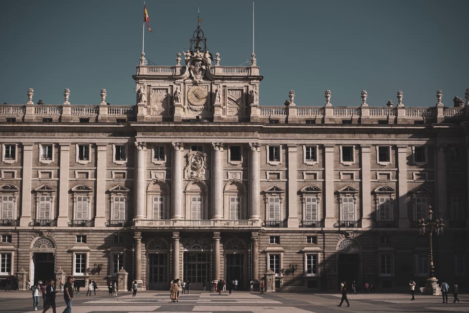 The middle of the palace front, close: columns, shuttered windows and a clock under a Spanish flag, with people small at the foot of it.