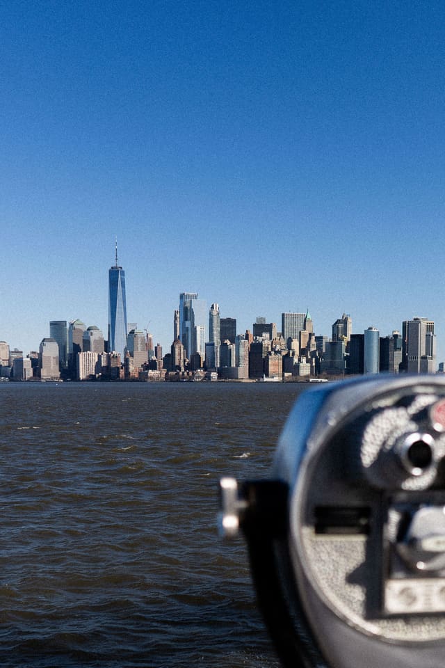 Lower Manhattan across the harbour under a clear sky, with a coin-operated viewer out of focus in the foreground.