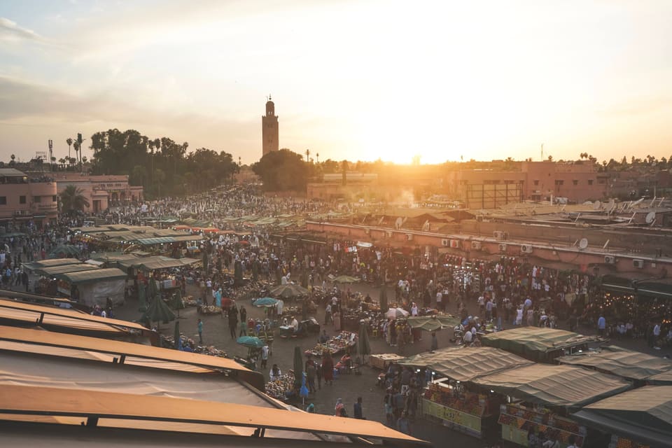 A crowded market square at sunset, seen from a rooftop: canvas stalls, hundreds of people, and a minaret against the low sun.