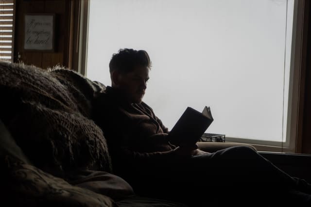 Alex in silhouette against a bright window, reading a book on a sofa.