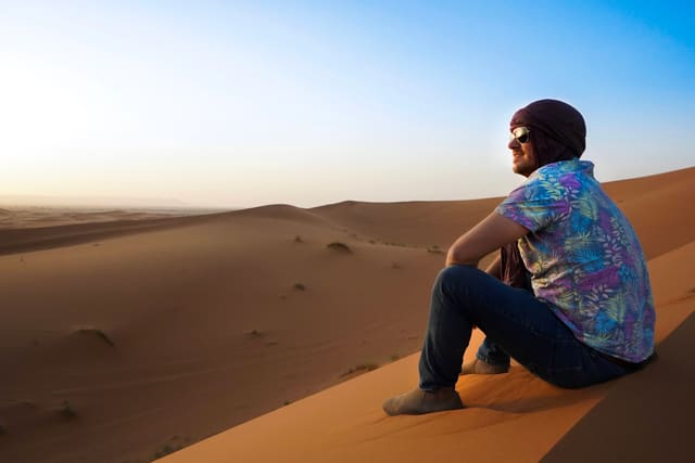 Alex sits on the crest of an orange sand dune in a head wrap and sunglasses, looking out over the desert in low sun.