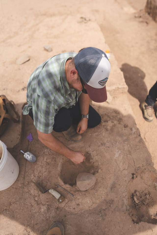 Seen from above, an archaeologist in a plaid shirt and cap kneels at a small round pit and reaches in beside a smooth stone, with a trowel and a scoop on the ground next to him.