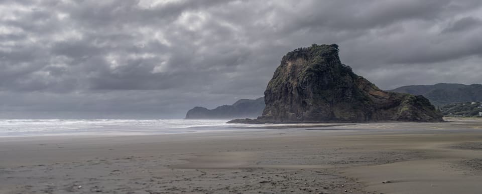A dark rock headland rising from a wide, wet beach under heavy grey cloud.