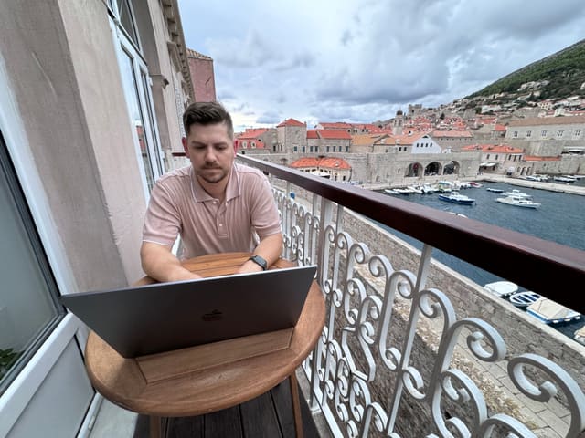 Alex at a laptop on a small round table on a balcony, with the old harbour, its boats and the terracotta roofs of Dubrovnik behind him.