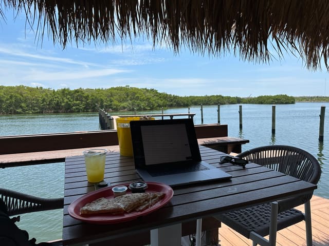 A laptop on a table under a thatched roof at the water’s edge, with lunch beside it and mangroves across the channel.