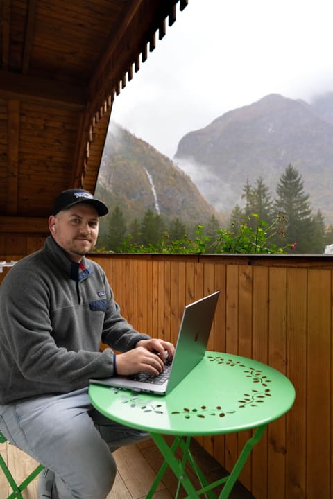 Alex at a laptop on a green café table on a wooden balcony, with a waterfall and mountains in low cloud behind him.