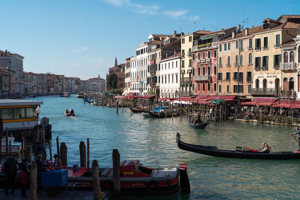 The Grand Canal in morning light: gondolas crossing, a water-bus stop, and restaurant awnings along the bank.