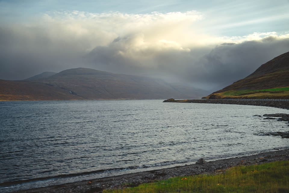 Storm light breaking over a fjord: dark water, a curving shoreline, and cloud lit from behind above the far hills.